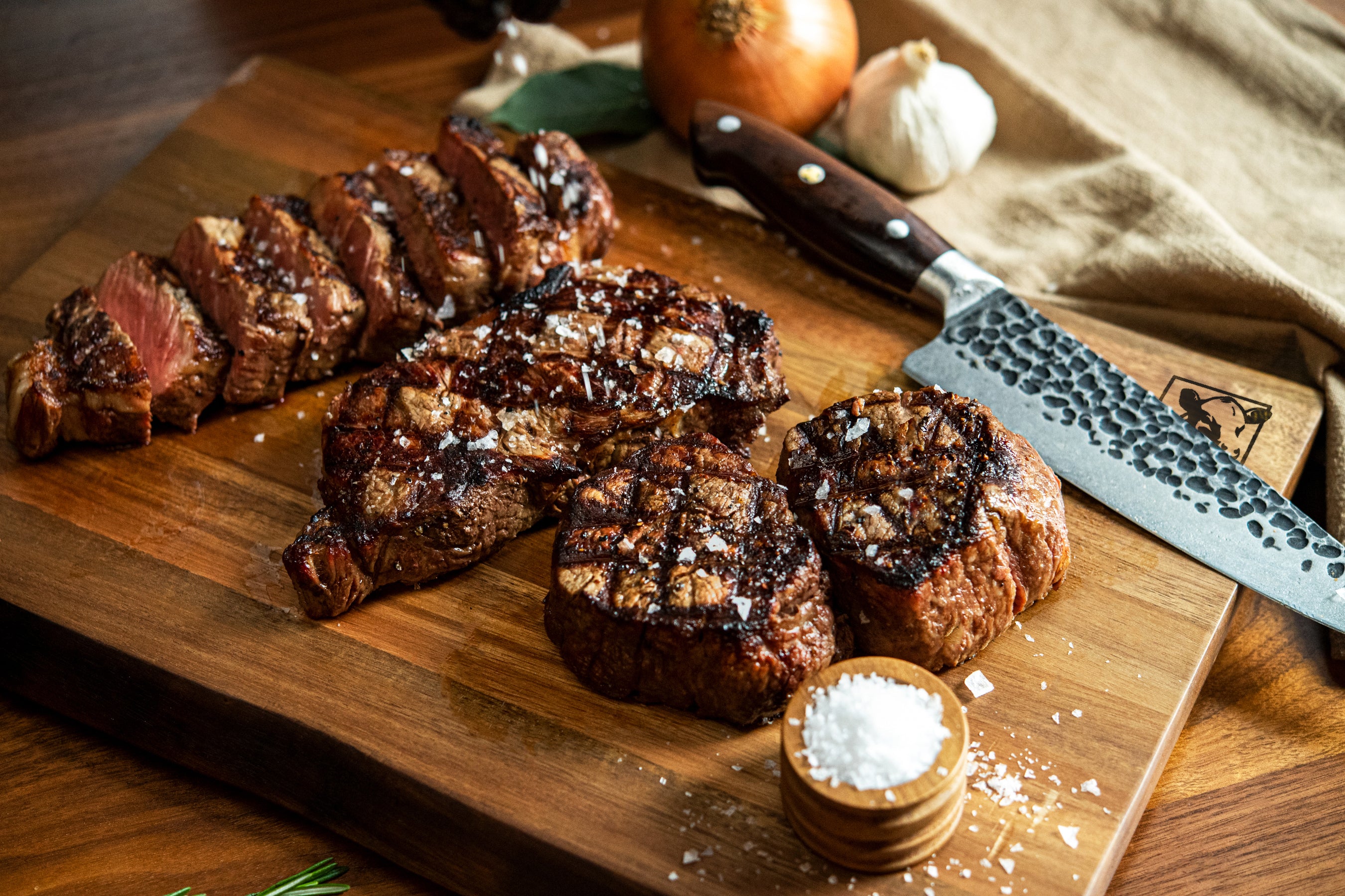 Sliced beef on a wooden cutting board with a knife, salt, and garlic.